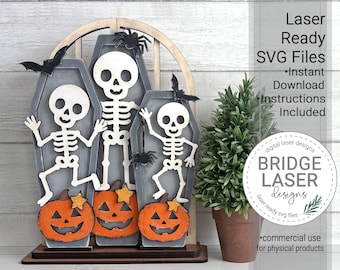 May include: A laser-cut wooden display featuring three grey coffins, each containing a white skeleton figure. Three orange jack-o'-lanterns with yellow stars sit at the base. Black spiders and bats are attached to the coffins. Text reads Laser Ready SVG Files, Instant Download, Instructions Included, and BRIDGE LASER designs.