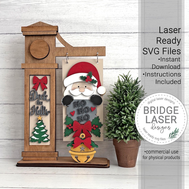 May include: A wooden laser-cut display stand featuring a vertical post with a red bow, the text Deck the Halls, and a green Christmas tree. A hanging sign shows Santa Claus with the text HO HO HO, a red bow, and a yellow bell at the bottom.