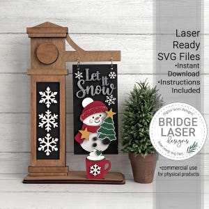 K&ouml;nnte beinhalten: Ein Holzschild-Display mit einer h&auml;ngenden schwarzen rechteckigen Tafel mit dem Text Let it Snow, einer Schneemann-Illustration und einer roten Tasse. Die Struktur umfasst einen braunen Holzpfosten, wei&szlig;e Schneeflocken und eine kleine gr&uuml;ne Pflanze.