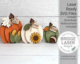 May include: Three wooden pumpkin cutouts in orange, white, and green. The orange pumpkin features corn and a leaf with a tag reading Hello Fall. The white pumpkin has a sunflower and a tag reading Gather. The green pumpkin has an acorn and a tag reading Blessed.
