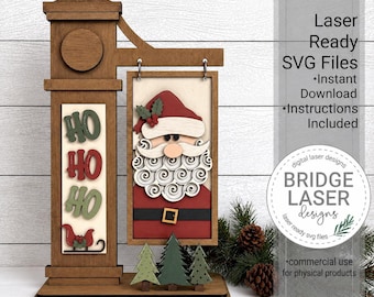 May include: A wooden laser-cut sign featuring a Santa Claus face and the text HO HO HO. The sign includes a vertical post, a hanging rectangular panel, and three small evergreen trees at the base. Text on the right reads Laser Ready SVG Files.