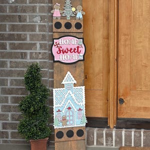 Könnte beinhalten: Ein Holzschild mit Lebkuchenhaus- und Lebkuchenmännchen-Design. Das Schild trägt die Worte "HOME SWEET HOME" in Rosa und Weiß. Es ist mit einem Weihnachtsbaum und einem Stern verziert.
