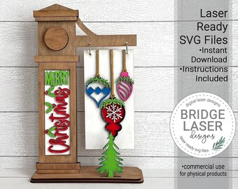 May include: Laser cut wooden Christmas sign with a hanging ornament display. The sign says "Merry Christmas" and features a red, green, blue, and pink ornament. The sign is on a white background.