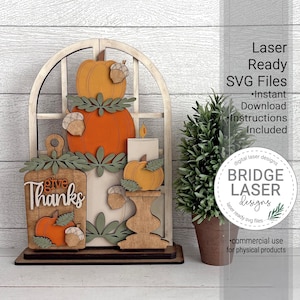 May include: A layered wooden laser-cut sign featuring an arched window frame, stacked orange and yellow pumpkins, acorns, and a cutting board shape with the text "give Thanks". A small potted green plant sits next to the decorative wooden display.