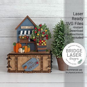Puede incluir: Un expositor de puesto de granja cortado con láser que incluye un edificio azul con toldo a rayas, calabazas y un manzano. La base de madera rectangular tiene una etiqueta azul con el texto FALL HARVEST. Incluye: Laser Ready SVG Files.