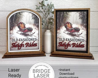 May include: Two wooden signs with the text "OLD FASHIONED Sleigh Rides" in red script. The signs feature a winter scene with a sleigh. One sign has an arched top, the other is rectangular. A vase with greenery is between the signs.