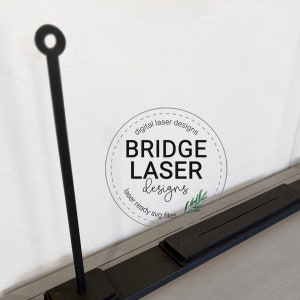 May include: Black metal laser cut design with a circular top and a rectangular base. The design includes the text "Bridge Laser Designs" in a circular logo.