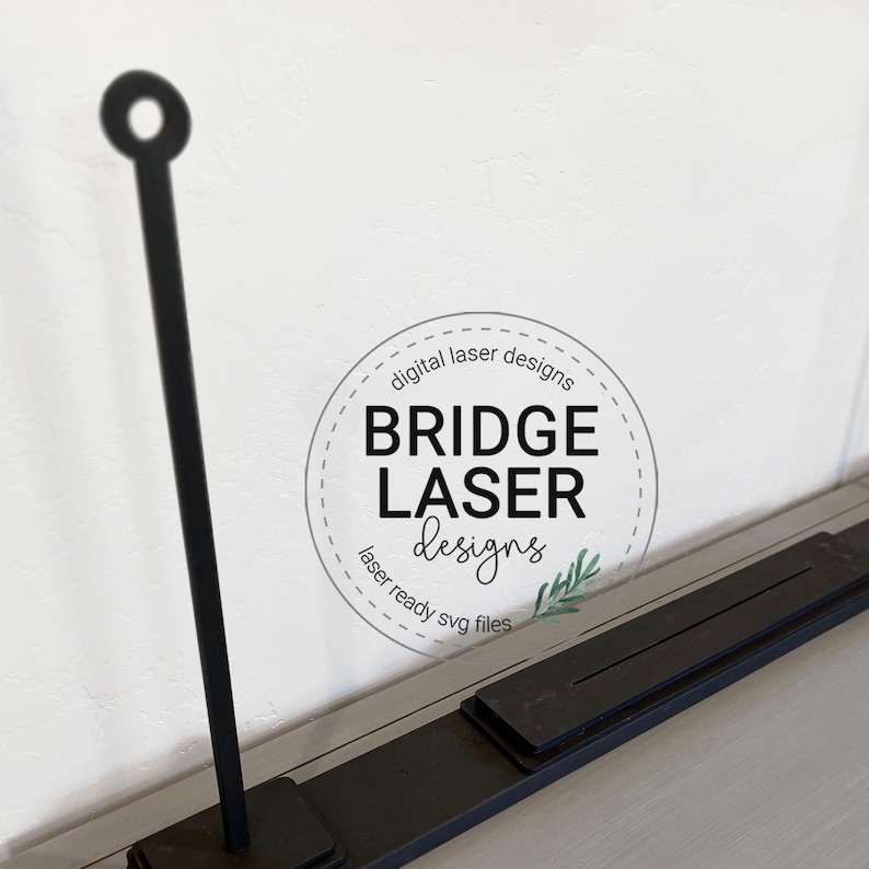 May include: Black metal laser cut design with a circular top and a rectangular base. The design includes the text "Bridge Laser Designs" in a circular logo.