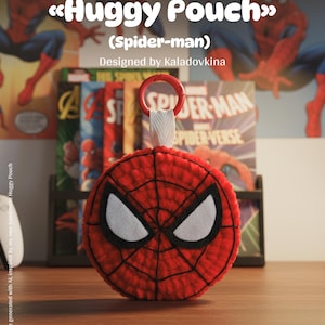 Op de afbeelding: Een rood en zwart gehaakt tasje in de vorm van Spider-Man's masker, met witte oogdetails. Het tasje heeft een rode ring en een witte band. De afbeelding bevat de tekst "Huggy Pouch" en "Spider-man".
