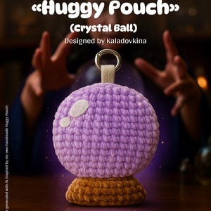 Op de afbeelding: Een gehaakte kristallen bol etui in tinten paars en bruin. De bolvormige etui heeft een getextureerd oppervlak en is bevestigd aan een metalen ring. De basis is donkerbruin van kleur. De tekst "Huggy Pouch" en "Crystal Ball" zijn zichtbaar.