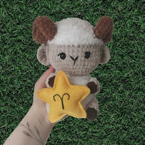 May include: A crocheted ram plushie with brown horns and hooves, and a white head. It holds a yellow star-shaped felt with the Aries symbol. The background is green.