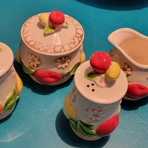 May include: A set of four white ceramic kitchenware items with floral and fruit designs. The set includes a creamer, a sugar bowl, a salt shaker, and a pepper shaker. Each piece features a different fruit design, including apples, pears, and oranges.