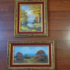 May include: Two framed landscape paintings. The top painting depicts a lake scene with yellow and green trees, a blue sky, and a mountain in the background. The bottom painting shows a field with trees and a small building under a blue sky.