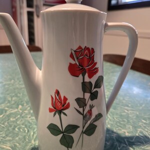 May include: A white porcelain coffee pot featuring a tall, cylindrical body with a matching lid topped by a spherical knob. The exterior is decorated with a graphic illustration of two red roses with green leaves and stems on the front surface.