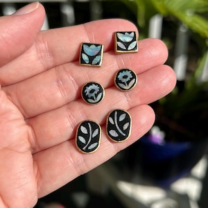 May include: A collection of six small stud earrings with gold-colored borders. The earrings feature black backgrounds with floral and leaf designs in white and iridescent blue. The earrings are in square, oval, and round shapes.