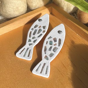 May include: Two white fish-shaped decorative objects with mosaic-style inlays of various colors. The fish have small, dark eye details and are displayed on a wooden tray. The background includes soft, off-white felted wool balls.