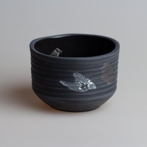 May include: A black ceramic bowl with a textured, ridged exterior. The bowl features abstract white and gray patterns on the side. The interior is also black. The bowl is likely handmade and suitable for serving food.