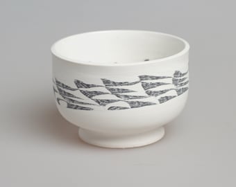 May include: A white ceramic cup with a black and white geometric pattern around the rim. The cup has a wide base and a rounded shape.