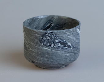 May include: A gray ceramic bowl with a marbled design. The bowl has a rounded shape with a slightly flared rim. The gray color is swirled with darker gray and white patterns, creating a unique, textured appearance. The bowl is suitable for serving food or as a decorative item.