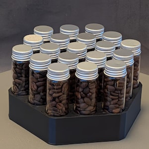 May include: A black hexagonal display stand holds 16 small glass vials filled with coffee beans. Each vial has a silver cap. The coffee beans are a dark brown color. The stand is on a neutral surface.