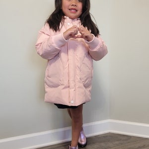 Puede incluir: Una niña pequeña con una chaqueta acolchada rosa con capucha haciendo un corazón con las manos. Lleva una falda negra y botas moradas.