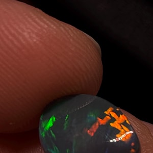 May include: A round, dark-toned opal gemstone featuring vibrant flashes of neon green and bright orange play-of-colour across its surface. The stone displays a smooth, polished finish with distinct colour patterns that shift against the dark base.