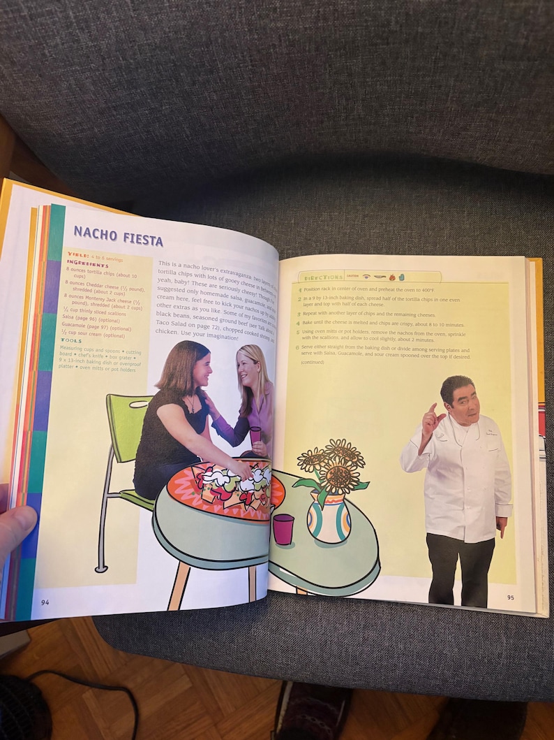 May include: A cookbook open at a double-page spread, with the title "NACHO FIESTA." The left page has an illustration of a table with food and two women. The right page shows a chef gesturing near a table with sunflowers.