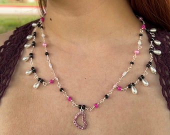 Puede incluir: Un collar de dos capas con cuentas negras y blancas, acentos rosados y un dije de corazón rosa. El collar se combina con aretes colgantes que presentan un dije de araña plateada y cuentas rosadas y negras.