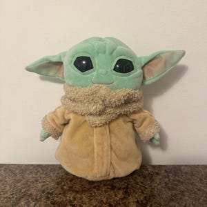 May include: A soft toy of a character from a popular science fiction series. The toy has large black eyes, large ears, and a light green face. It is wearing a tan coat and a fluffy, tan scarf.