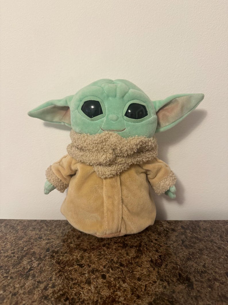 May include: A soft toy of a character from a popular science fiction series. The toy has large black eyes, large ears, and a light green face. It is wearing a tan coat and a fluffy, tan scarf.
