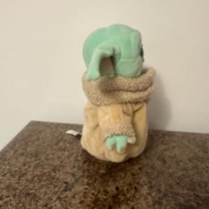May include: A plush toy of a character with large ears and green skin, wearing a tan coat with a furry collar and cuffs. The toy is sitting on a brown surface.