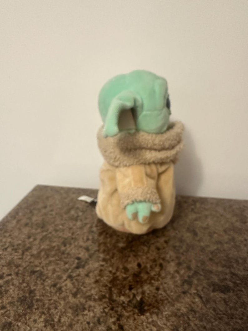 May include: A plush toy of a character with large ears and green skin, wearing a tan coat with a furry collar and cuffs. The toy is sitting on a brown surface.
