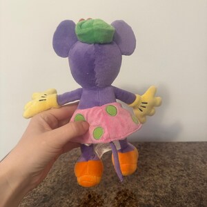 Pu&ograve; includere: Un peluche a forma di topo con corpo viola, grandi orecchie e cappello verde. Ha una gonna rosa a pois verdi, scarpe arancioni e guanti gialli. Il giocattolo &egrave; tenuto da una persona.
