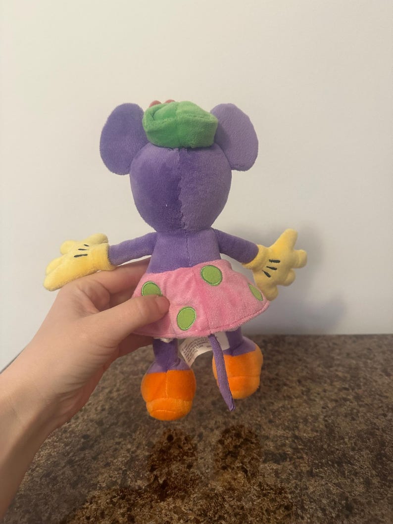Pu&ograve; includere: Un peluche a forma di topo con corpo viola, grandi orecchie e cappello verde. Ha una gonna rosa a pois verdi, scarpe arancioni e guanti gialli. Il giocattolo &egrave; tenuto da una persona.