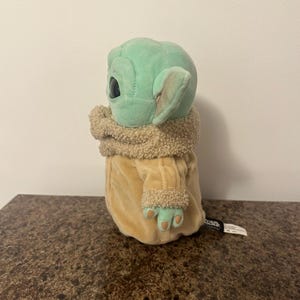 May include: A plush toy of a character from a popular science fiction series. The toy has a light green head with large black eyes and pointed ears. It wears a tan coat with a fuzzy collar and cuffs. The toy is sitting on a brown surface.