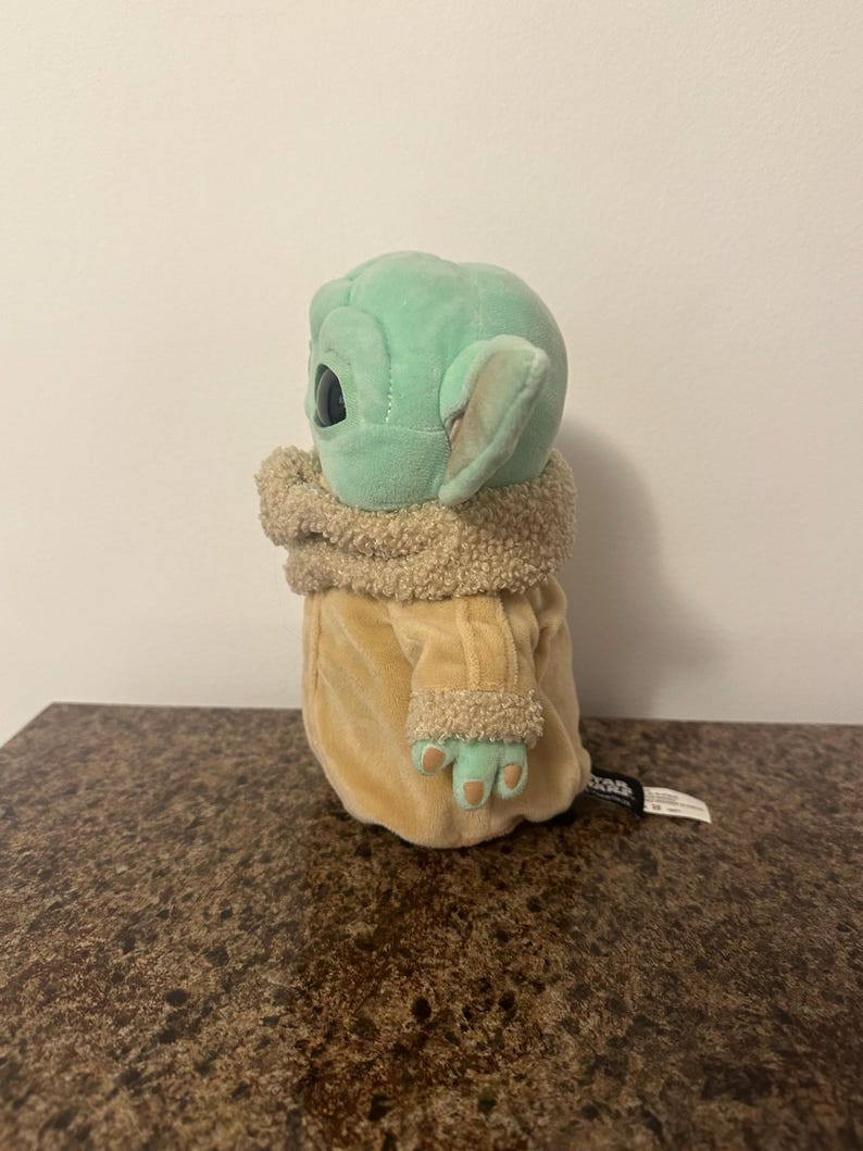 May include: A plush toy of a character from a popular science fiction series. The toy has a light green head with large black eyes and pointed ears. It wears a tan coat with a fuzzy collar and cuffs. The toy is sitting on a brown surface.