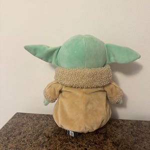 May include: A soft toy of a character from the Star Wars franchise, with a light green head and large ears, a tan coat, and a fluffy collar. The toy is on a brown surface.