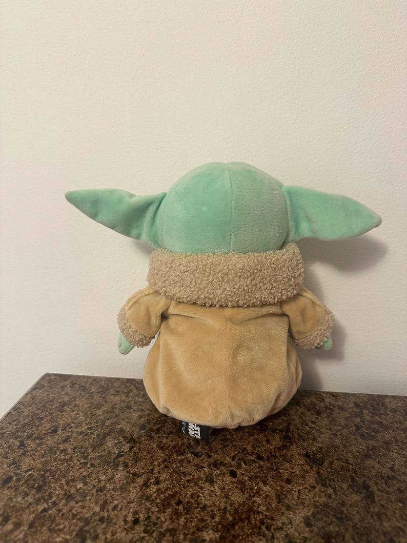 May include: A soft toy of a character from the Star Wars franchise, with a light green head and large ears, a tan coat, and a fluffy collar. The toy is on a brown surface.