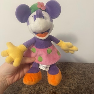 Pu&ograve; includere: Una bambola di peluche di Minnie Mouse con orecchie, braccia e gambe viola, viso bianco e gonna rosa a pois verdi. Indossa un fiore sulla testa e guanti gialli. La bambola &egrave; di Disney Parks.