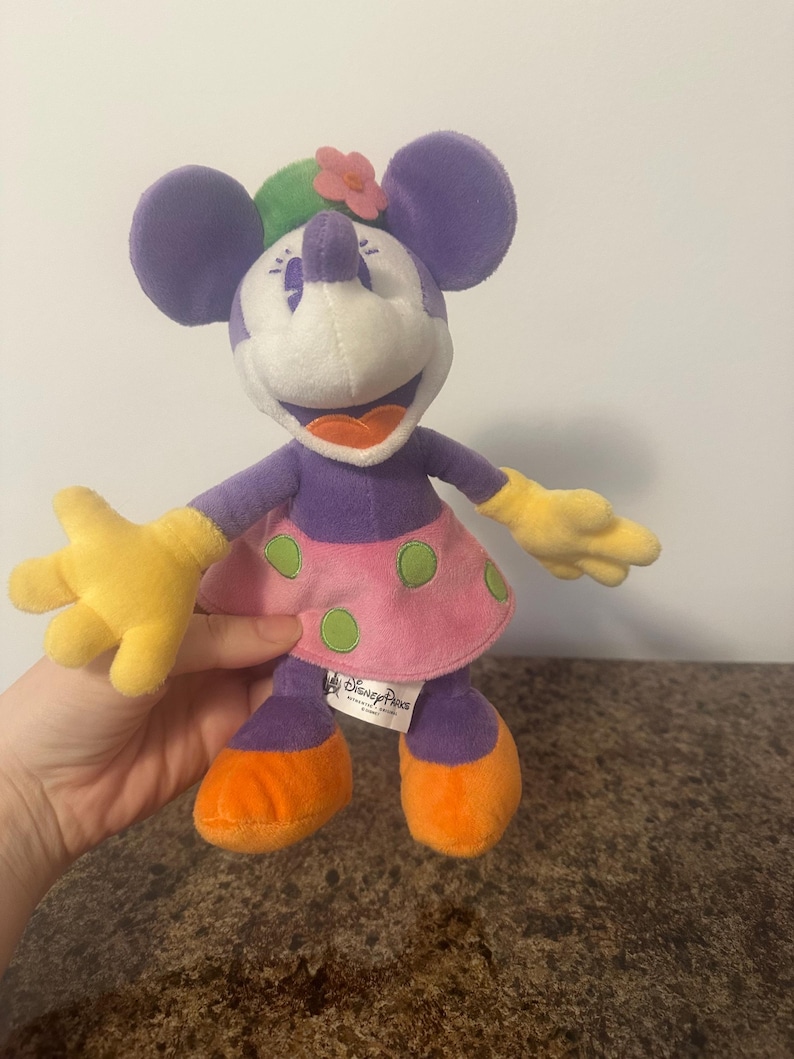 Pu&ograve; includere: Una bambola di peluche di Minnie Mouse con orecchie, braccia e gambe viola, viso bianco e gonna rosa a pois verdi. Indossa un fiore sulla testa e guanti gialli. La bambola &egrave; di Disney Parks.
