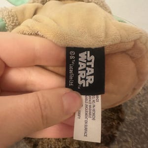 May include: A tan plush toy of a Star Wars character with green accents and a fuzzy head. A black tag with the Star Wars logo and text is visible. The tag also includes washing instructions.