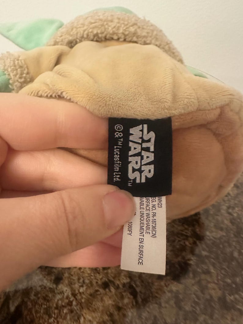 May include: A tan plush toy of a Star Wars character with green accents and a fuzzy head. A black tag with the Star Wars logo and text is visible. The tag also includes washing instructions.