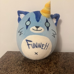 May include: A plush toy cat with a blue body, blue ears, and a yellow and orange striped horn. The cat has a white belly with the word "FUNNEH" embroidered in blue, and a small black "x" below the word. The cat has black markings around the eyes.