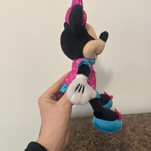 May include: A plush Minnie Mouse doll, featuring a black head and ears, a pink polka-dot dress, and white gloves. The doll has a pink bow and blue accents. The shoes are black with blue soles and red bows.
