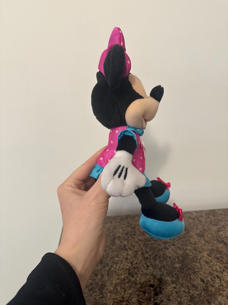 May include: A plush Minnie Mouse doll, featuring a black head and ears, a pink polka-dot dress, and white gloves. The doll has a pink bow and blue accents. The shoes are black with blue soles and red bows.