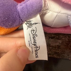 Pu&ograve; includere: Primo piano di un peluche con un braccio viola e un vestito rosa. &Egrave; visibile un'etichetta bianca con il logo Disney Parks e le parole "AUTHENTIC ORIGINAL".