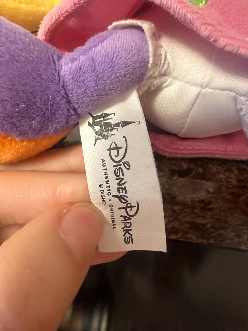 Pu&ograve; includere: Primo piano di un peluche con un braccio viola e un vestito rosa. &Egrave; visibile un'etichetta bianca con il logo Disney Parks e le parole "AUTHENTIC ORIGINAL".