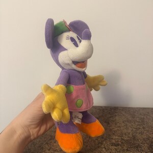 Pu&ograve; includere: Bambola di peluche di Minnie Mouse. La bambola ha testa e corpo viola, viso bianco, mani gialle e scarpe arancioni. Indossa un vestito rosa a pois verdi e un cappello verde e rosa. Il logo Disney &egrave; visibile.