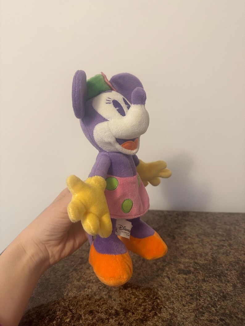 Pu&ograve; includere: Bambola di peluche di Minnie Mouse. La bambola ha testa e corpo viola, viso bianco, mani gialle e scarpe arancioni. Indossa un vestito rosa a pois verdi e un cappello verde e rosa. Il logo Disney &egrave; visibile.
