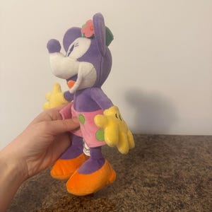 Pu&ograve; includere: Bambola di peluche di Minnie Mouse in un abito tropicale. La bambola ha testa e corpo viola, viso bianco, guanti gialli, scarpe arancioni e gonna rosa a pois verdi. Un fiore rosa e una foglia verde adornano la sua testa.